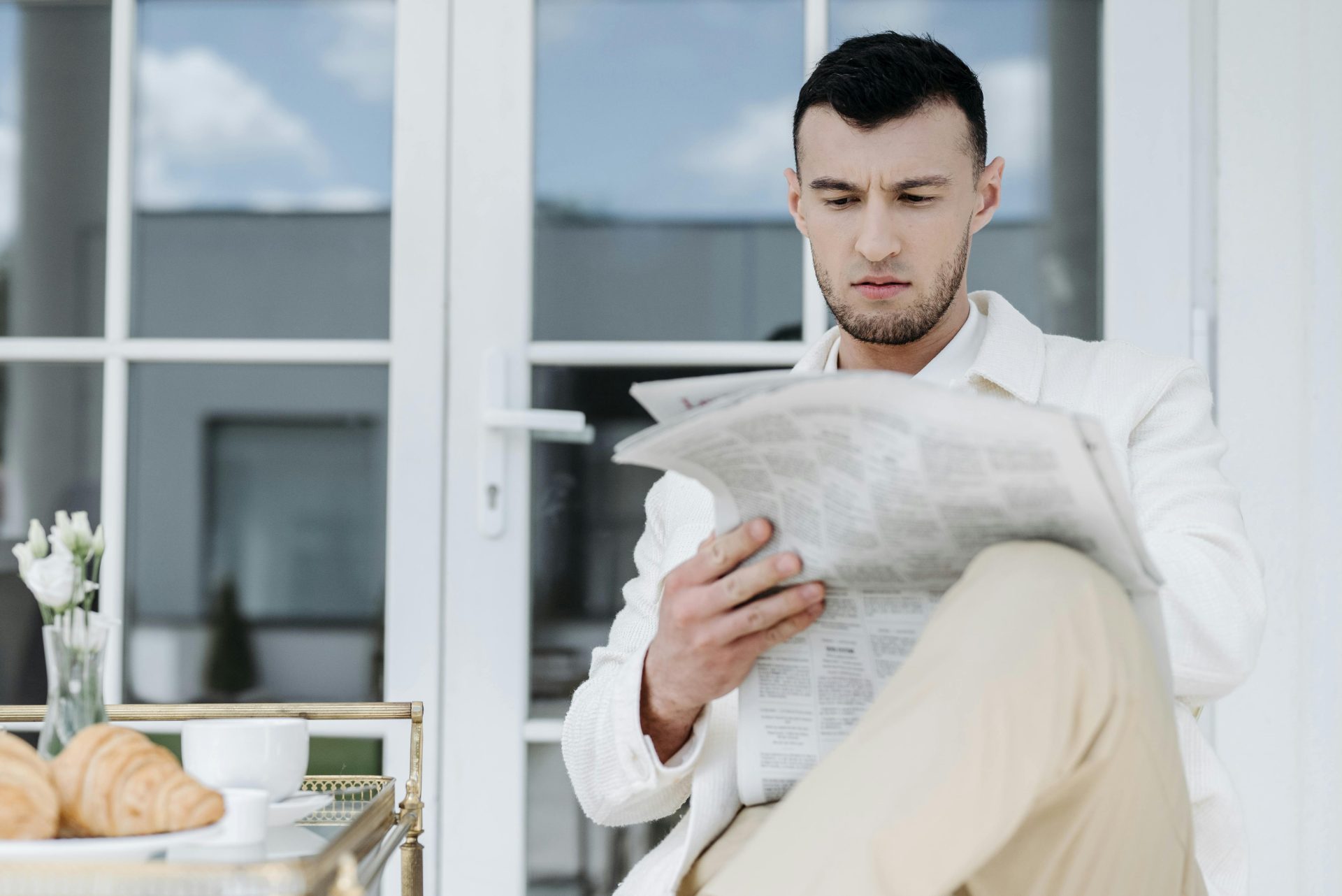 A man in white attire reads a newspaper outdoors, enjoying a breakfast with croissants and coffee.